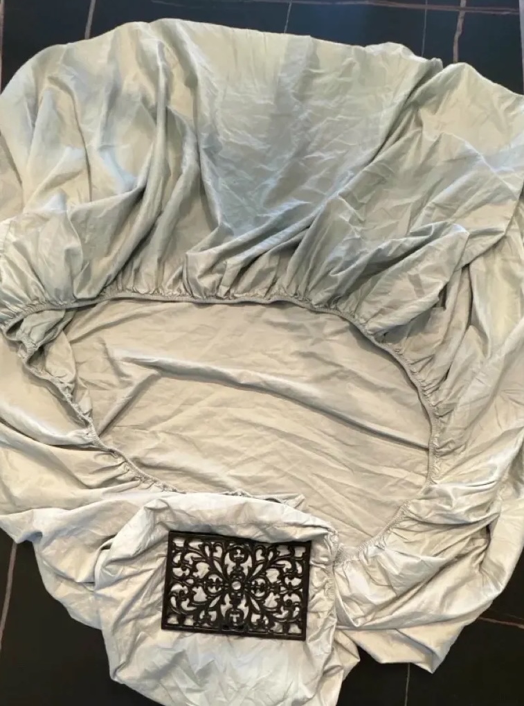 Cast iron trivet holding down nested fitted sheet corners on a dark dining table, blue pants and tile floor visible